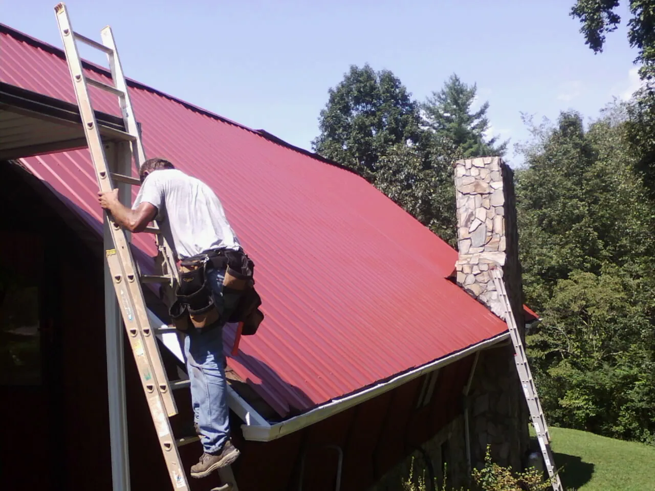 Roofer on a ladder beside a red metal roof and stone chimney — Soffit & Fascia Repair by Roofing Brothers in Blowing Rock